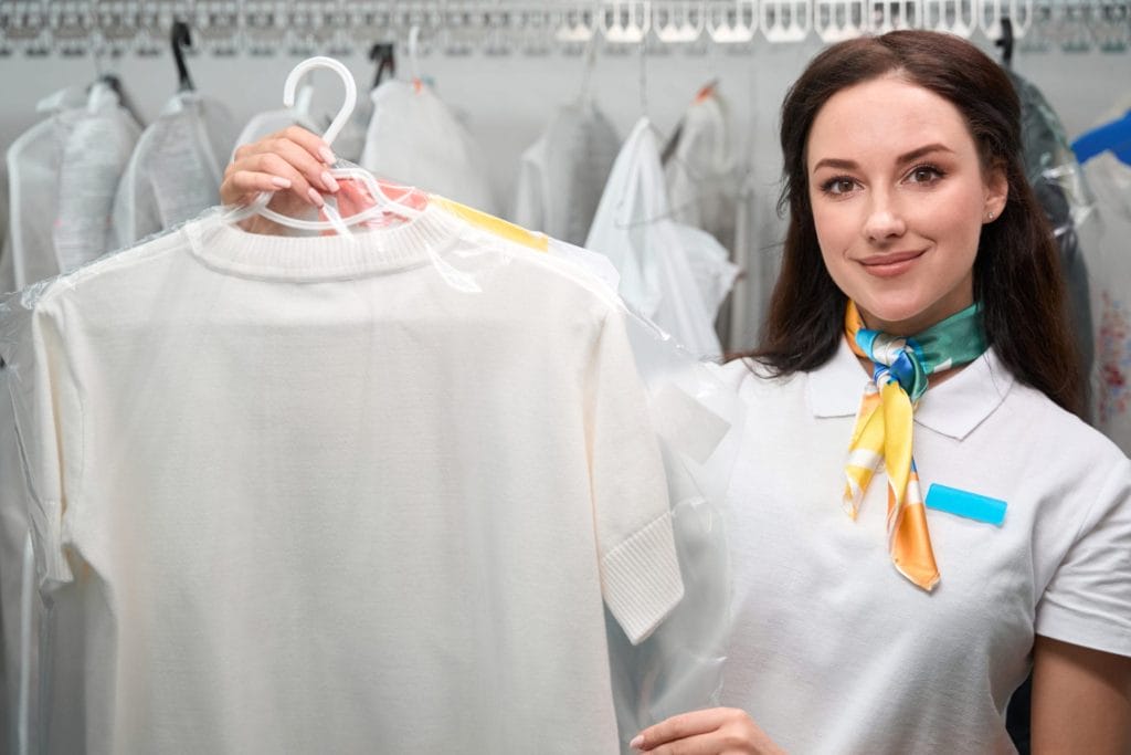 A female employee in a dry cleaning shop, wearing a uniform with a yellow scarf, holds up a white shirt on a hanger, smiling at the camera.